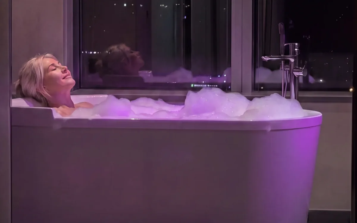 A woman lying back in a bath full of bubbles lit violet