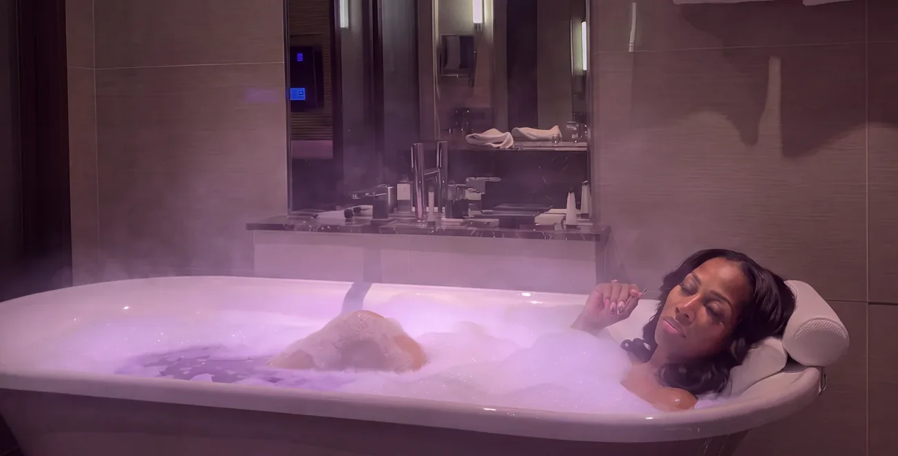 A woman lying back in a deep bath of violet lit bubbles, steam rising around her